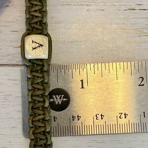 K&ouml;nnte beinhalten: Ein gr&uuml;nes Lederarmband mit einer wei&szlig;en quadratischen Perle, auf der der Buchstabe "V" steht. Das Armband wird mit einem Lineal gemessen und ist etwa 2,5 cm breit.