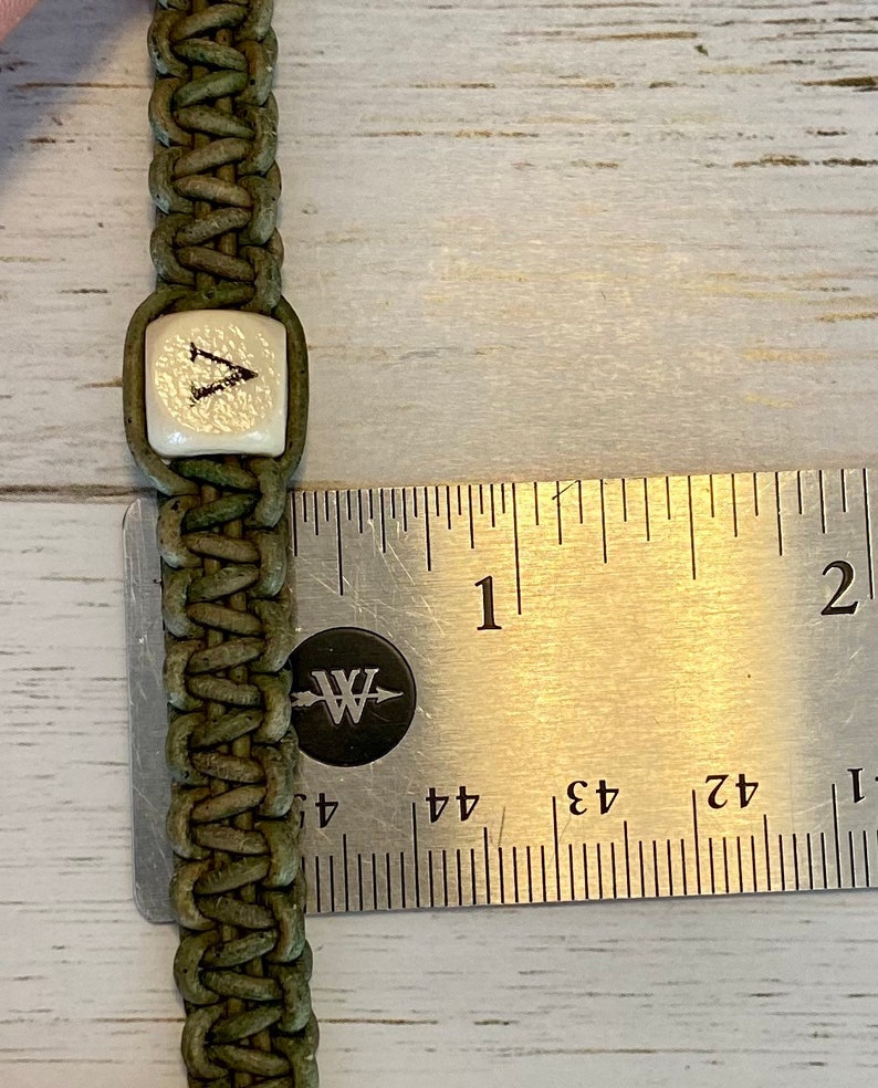 K&ouml;nnte beinhalten: Ein gr&uuml;nes Lederarmband mit einer wei&szlig;en quadratischen Perle, auf der der Buchstabe "V" steht. Das Armband wird mit einem Lineal gemessen und ist etwa 2,5 cm breit.