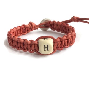 May include: A coral-colored macrame bracelet with a wooden bead featuring the letter "H". The bracelet has a button closure and leather cord details. The bracelet is a handmade accessory.