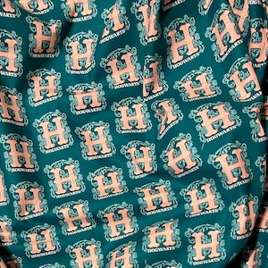 May include: A teal blue fabric with a repeating pattern of the Hogwarts crest. The crest is a shield with a lion, eagle, badger, and snake. The text "HOGWARTS" is written in a white script font.
