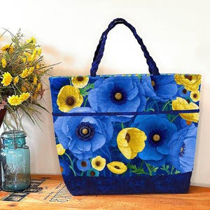 May include: A blue tote bag with a floral pattern of large blue and yellow flowers. The bag has a braided blue handle and a dark blue trim. The background includes a vase of yellow flowers and a blue glass jar.