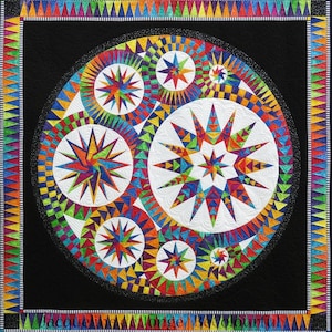 May include: A colorful quilt with a black background and a circular design featuring starbursts and geometric patterns in vibrant colors. The quilt has a border with triangles and the text "Jacqueline de Jonge Beautiful".