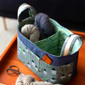 May include: A fabric yarn storage basket with a floral pattern in gray, white, and green. The basket has a denim blue trim and handles. Inside are two balls of yarn in gray and light gray. A pair of scissors and more yarn are in front.