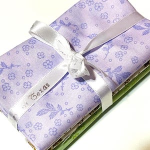 May include: A stack of folded fabric pieces tied with a white ribbon. The top layer is light purple with a dark purple floral and leaf pattern. The ribbon features the text "Texas" printed in a dark font. The lower fabric layers are green and white.