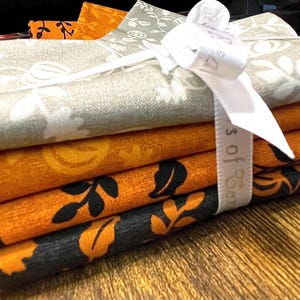 May include: A stack of four folded fabric squares tied together with a white ribbon. The top fabric is light gray with a white botanical pattern, followed by a solid orange fabric, an orange fabric with black leaves, and a black fabric with orange leaves.