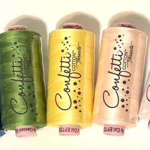 May include: A collection of seven spools of Confetti Cotton Threads in various colors, including navy, blue, green, yellow, peach, white, and black. Each spool has a pink base and the brand name printed on the side.