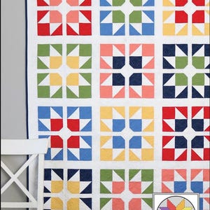 May include: A colorful quilt with a repeating split star pattern. The quilt features squares in various colors, including red, blue, green, and yellow, set against a white background. The text "Split Stars" is at the top. A white chair is in the left corner.