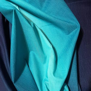 May include: Close-up of draped fabric in shades of blue and teal. The fabric has a textured appearance, with folds and shadows creating depth. The color gradient transitions from a deep navy to a vibrant teal, showcasing a smooth, flowing texture.