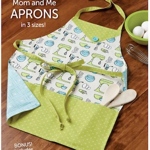 May include: A light green and white apron with a kitchen utensil pattern, featuring a matching green pocket and ties. The apron includes the text "Mom and Me APRONS in 3 sizes!" and "BONUS! includes Chenille Hotpad!" Wooden spoons are in the pocket.