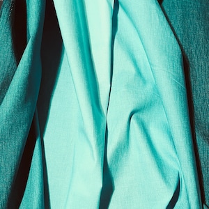 May include: Close-up of turquoise fabric with varying textures and folds. The fabric appears to be a blend of smooth and textured materials, creating depth and visual interest. The color is a vibrant teal, with shadows adding dimension.