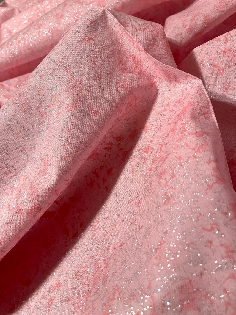 May include: Close-up of pink fabric with a marbled design. The fabric has a subtle shimmer and a textured appearance. The folds and shadows create depth and dimension, highlighting the fabric's soft texture.