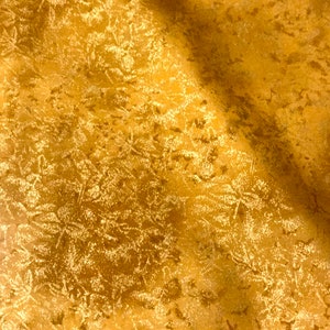 May include: Close-up of a golden fabric with a textured surface. The fabric has a pattern of small, light-colored floral or leaf-like shapes scattered across the surface. The fabric is folded, creating shadows and highlights.