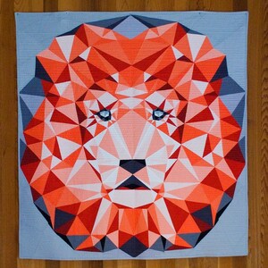 May include: Geometric lion quilt in shades of orange, red, white, and gray. The lion's face is constructed from triangular shapes, with blue eyes and a gray background. The quilt is square and appears to be handmade.