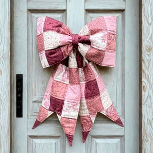May include: A large, quilted bow in shades of pink and burgundy hangs on a white door. The bow is made of patchwork squares with floral and geometric patterns. The bow's tails have pointed ends, adding a decorative touch to the door.