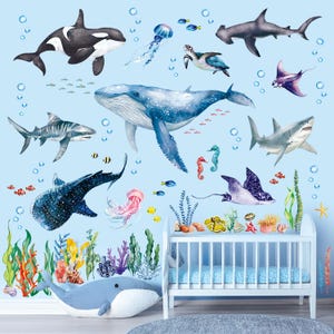 Pode incluir: Um conjunto de decalques de parede lúdicos com vários animais marinhos, incluindo uma baleia, tubarões e uma tartaruga, em tons de azul, cinza e branco. O design inclui bolhas e corais coloridos, ideal para um quarto infantil.