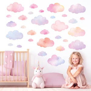 May include: Watercolor cloud wall decals in shades of pink, peach, and lavender. A crib with a pink blanket and a unicorn plush toy are in the foreground. A young girl in a pink dress is sitting on the floor.