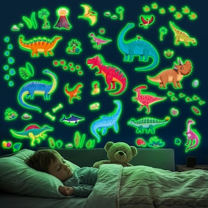 May include: A child asleep in bed with a glowing dinosaur wall decal. The decals are various colors, including green, orange, blue, and pink. The dinosaurs are surrounded by green glowing accents. A teddy bear is in the bed with the child.