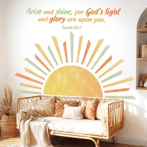 May include: A wall decal featuring a watercolor-style sun with rays in shades of yellow, orange, and green. The text "Arise and shine, for God's light and glory are upon you. Isaiah 60:1" is above the sun. A rattan daybed with pillows is below.