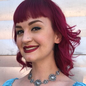 May include: A silver-toned necklace with circular and star-shaped pendants. The necklace is worn by a person with red hair and a blue top. The pendants have a detailed, vintage design. The person is smiling.