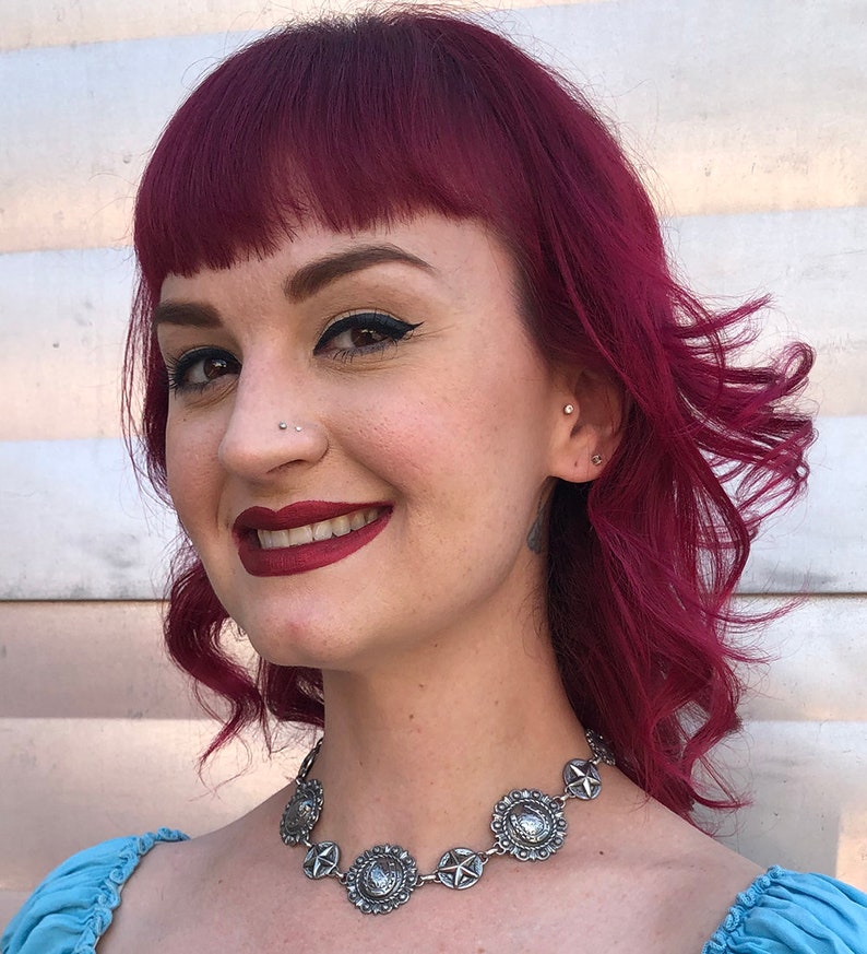 May include: A silver-toned necklace with circular and star-shaped pendants. The necklace is worn by a person with red hair and a blue top. The pendants have a detailed, vintage design. The person is smiling.