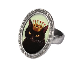 May include: A silver ring with an oval-shaped cameo featuring a black cat wearing a gold crown with red jewels. The cameo is set in a silver frame with a textured border.