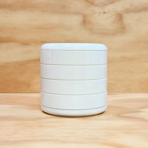 May include: A cylindrical white container featuring a stackable design with four distinct horizontal segments. The object has a smooth, solid white finish and a rounded top lid, presenting a clean and uniform appearance throughout its structure.