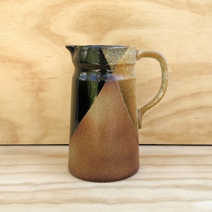 May include: A ceramic pitcher with a handle, featuring a geometric design. The pitcher has a black, tan, and brown color scheme. The pitcher is sitting on a wooden surface.