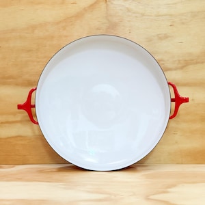 May include: A round, shallow cooking pan with a smooth, white enameled interior and a thin black rim. The pan features two bright red, curved metal handles attached to opposite sides, providing a clear contrast against the white basin.