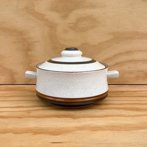 May include: A ceramic lidded casserole dish with a speckled white body, brown and black bands, and small side handles. The lid has a matching design with a black knob. The dish is set against a light wood grain background.