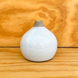 May include: A small, round ceramic vase with a glossy white glaze and a speckled finish. The vase features a narrow, tapered neck with a matte, light brown rim. The body of the vessel is bulbous and smooth, narrowing toward the top opening.