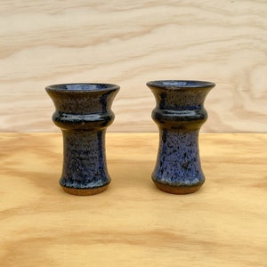 May include: Two handmade ceramic vases. The vases are dark blue with a speckled texture and a flared top. The base is a lighter brown color. The vases are displayed on a light wood surface.