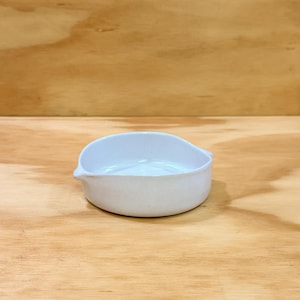 May include: A small, white ceramic bowl with a pouring spout. The bowl has a rounded shape with a slightly raised rim on one side. The interior of the bowl is smooth and white, and the exterior has a matte finish. The bowl is sitting on a wooden surface.