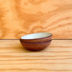 May include: Two small, brown ceramic bowls stacked on top of each other. The bowls have a rounded shape with a lighter-colored interior and a darker brown exterior. The bowls are set against a light brown wooden background.