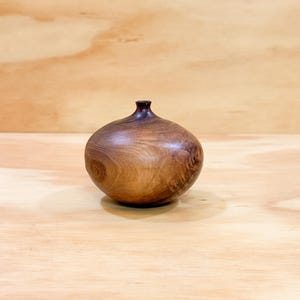 May include: A small, rounded wooden vase with a smooth, polished finish. The object features a bulbous body that tapers into a narrow, circular opening at the top. The wood displays a rich, dark brown grain pattern with visible natural swirls.