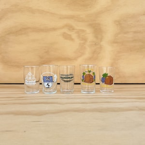 May include: Five clear shot glasses with various colorful designs. The designs include a crest, a bridge, and images of grapes and barrels. The glasses are arranged on a light brown wooden surface.