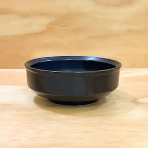 May include: A dark blue ceramic bowl with a rounded base and a slightly flared rim. The bowl is set against a light wood grain background, creating a contrast in textures and colors. The bowl's interior is a deep, solid blue.
