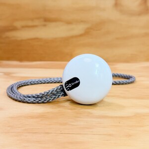 May include: A white, spherical Terraillon kitchen timer featuring a black display panel with white text and markings. The device is attached to a grey, braided cord that loops around the base of the round, glossy white plastic housing.