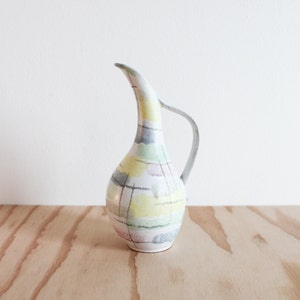 May include: A tall, slender ceramic pitcher with a curved handle. The pitcher is painted with a colorful, abstract pattern of yellow, green, gray, and pink blocks, overlaid with black lines. The pitcher sits on a light wood surface.