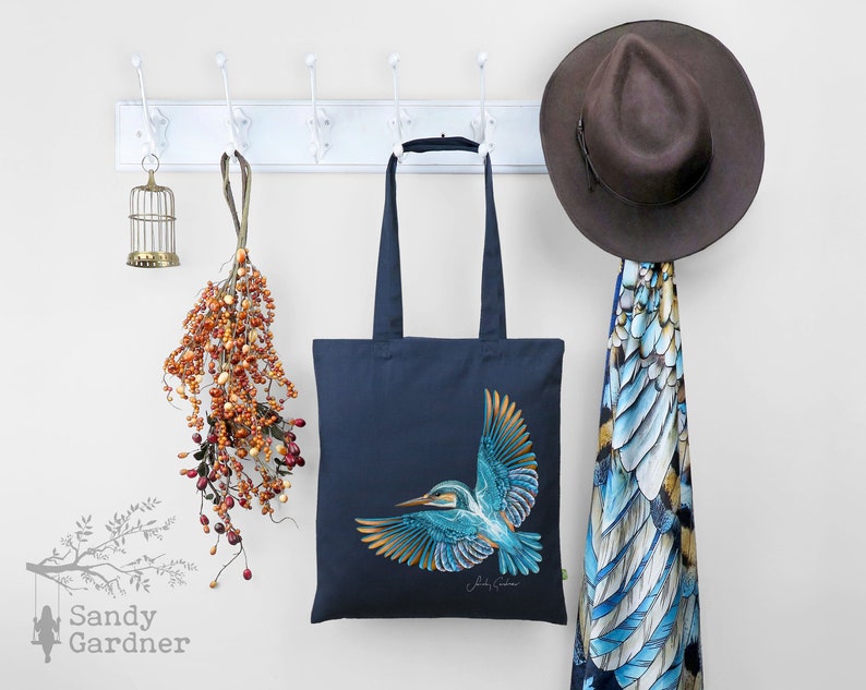 K&ouml;nnte beinhalten: Eine marineblaue Tragetasche mit einer bunten Illustration eines Eisvogels. Die Tasche hat einen langen Henkel und h&auml;ngt an einem wei&szlig;en Garderobenst&auml;nder. Ein brauner Hut und ein blau-goldener Schal h&auml;ngen ebenfalls am St&auml;nder.