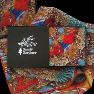 May include: A black box with a white logo that says "Sandy Gardner" and a small image of a girl on a swing. The box is open and shows a colorful silk pocket square with a pheasant design. The pocket square is folded and has a red, orange, and brown color scheme.