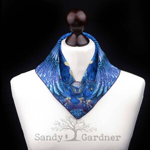 May include: A blue and purple patterned silk scarf with a large, round, blue gemstone brooch. The scarf is folded into a collar shape and is displayed on a white mannequin.