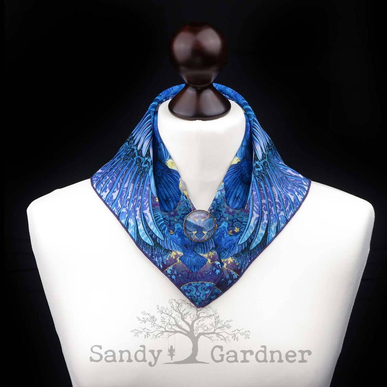 May include: A blue and purple patterned silk scarf with a large, round, blue gemstone brooch. The scarf is folded into a collar shape and is displayed on a white mannequin.