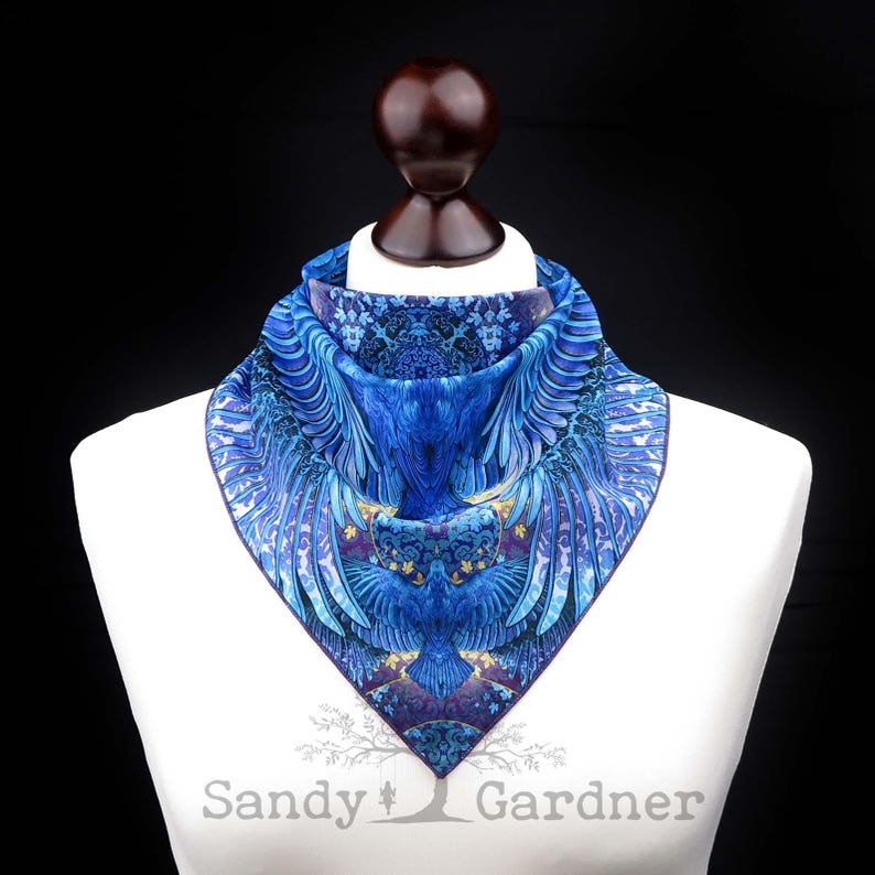 May include: A blue and purple silk scarf with a detailed design of a bird with outstretched wings. The scarf is displayed on a white mannequin bust.