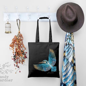 K&ouml;nnte beinhalten: Schwarze Einkaufstasche mit einer farbenfrohen Illustration eines Eisvogels im Flug. Die Tasche hat einen wei&szlig;en Henkel und h&auml;ngt an einem wei&szlig;en Garderobenst&auml;nder. Die Unterschrift des K&uuml;nstlers ist am unteren Rand der Illustration zu sehen.
