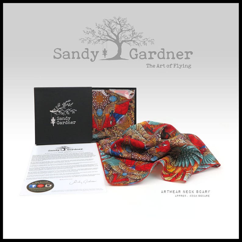 May include: A red, blue, and green silk neck scarf with a bird and floral pattern. The scarf is folded and displayed in a black box with the brand name "Sandy Gardner" and the text "The Art of Flying". The scarf is labeled "Artwear Neck Scarf Approx. 43cm Square".