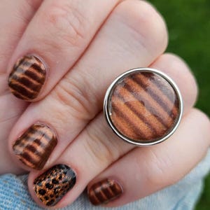May include: A silver ring with a round, brown and orange striped design. The ring is on a hand with brown and black nail polish with a striped and leopard print design. The background is a blurred green.