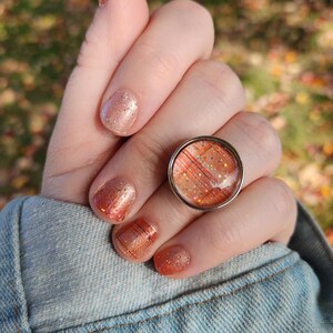 May include: A round silver ring with a plaid design in shades of orange, red, and white. The ring is worn on a hand with matching orange plaid nail polish. The background is blurred with fall colors.