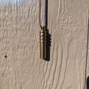 Pode incluir: Um pingente de metal dourado pendurado em um cordão roxo. O pingente é cilíndrico e tem uma série de anéis ao redor.