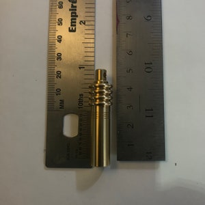May include: A brass-colored cylindrical object with a ridged section and a small top, next to a ruler. The object appears to be approximately 3 inches long. The ruler is marked with inches and tenths.