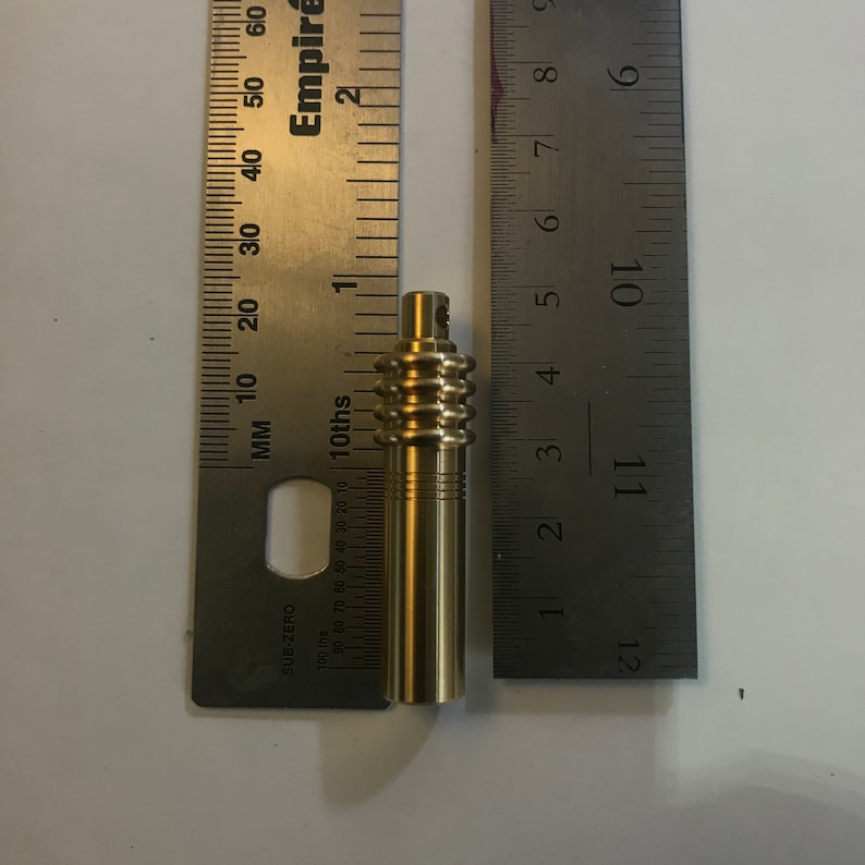 May include: A brass-colored cylindrical object with a ridged section and a small top, next to a ruler. The object appears to be approximately 3 inches long. The ruler is marked with inches and tenths.